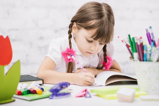Criança escrevendo em um caderno, com material escolar sobre a mesa.