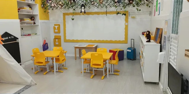 Sala de Educação Infantil montada, com mesas e cadeiras baixas, estante de livros e cantinho de leitura.